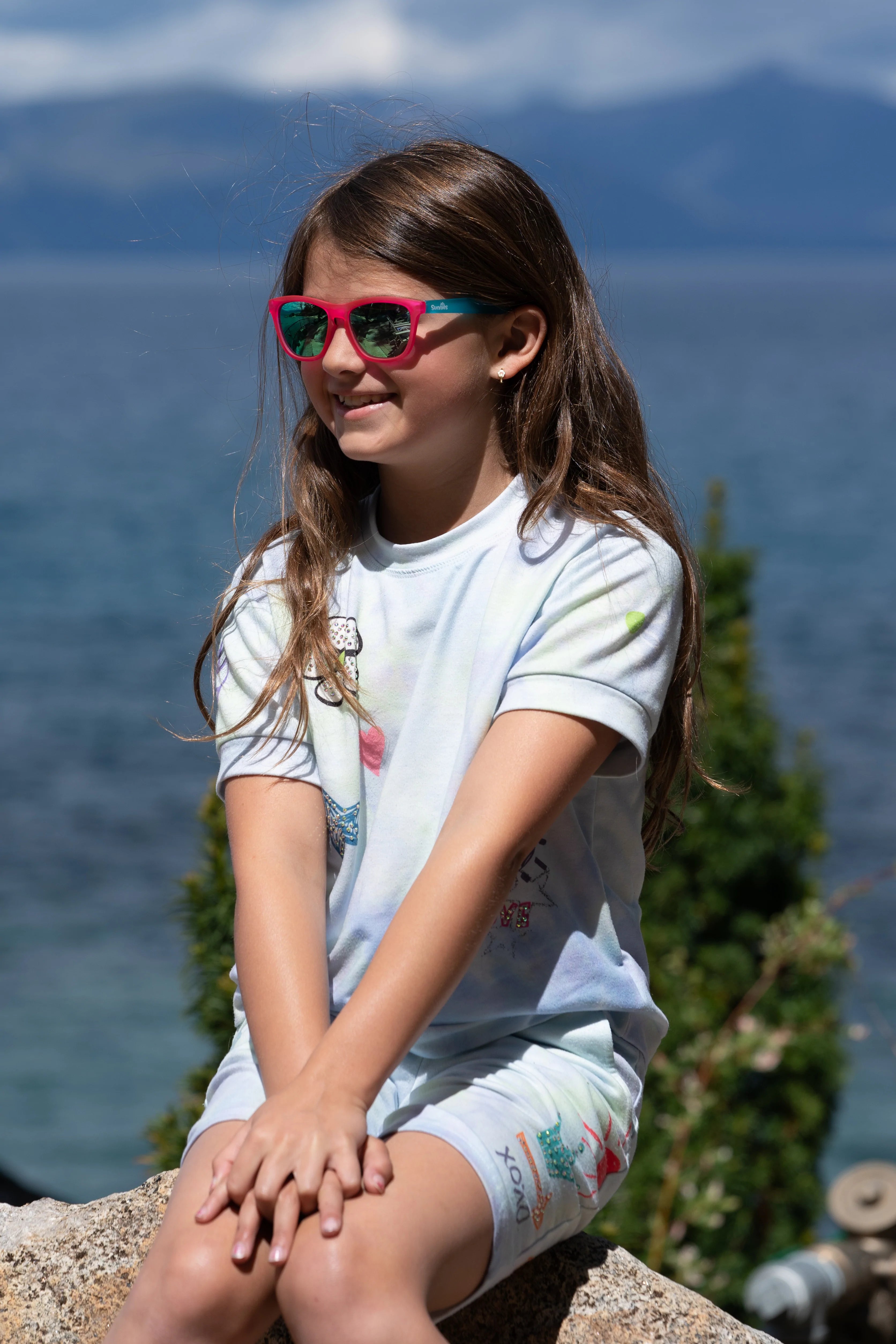 girl in sunglasses that are pink and turquoise sitting on a rock with a lake behind her