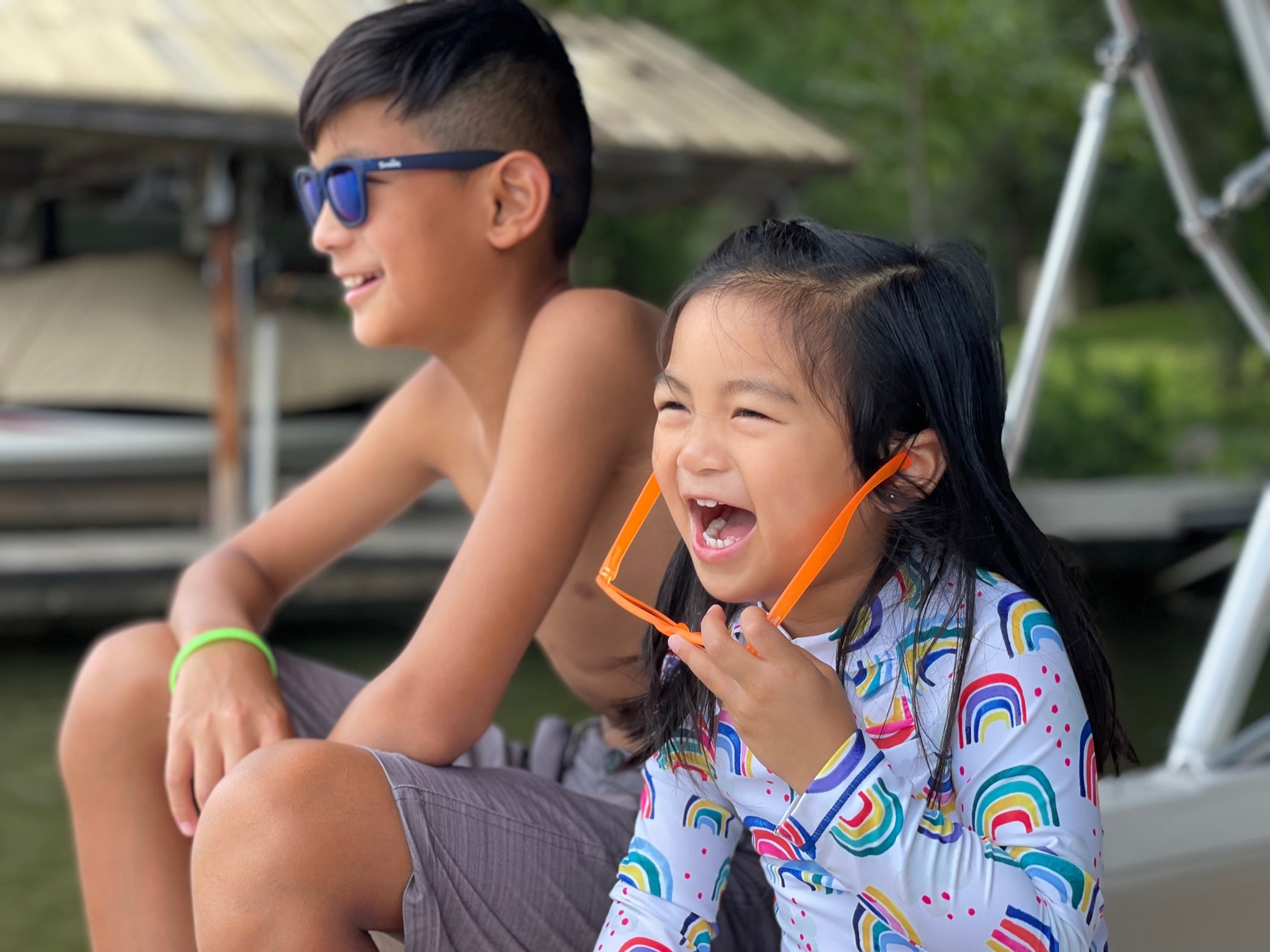 A boy and a girl sitting by a lake in bathing suits wearing Sunnies kids polarized sunglasses with 100% UVA/UVB protection.