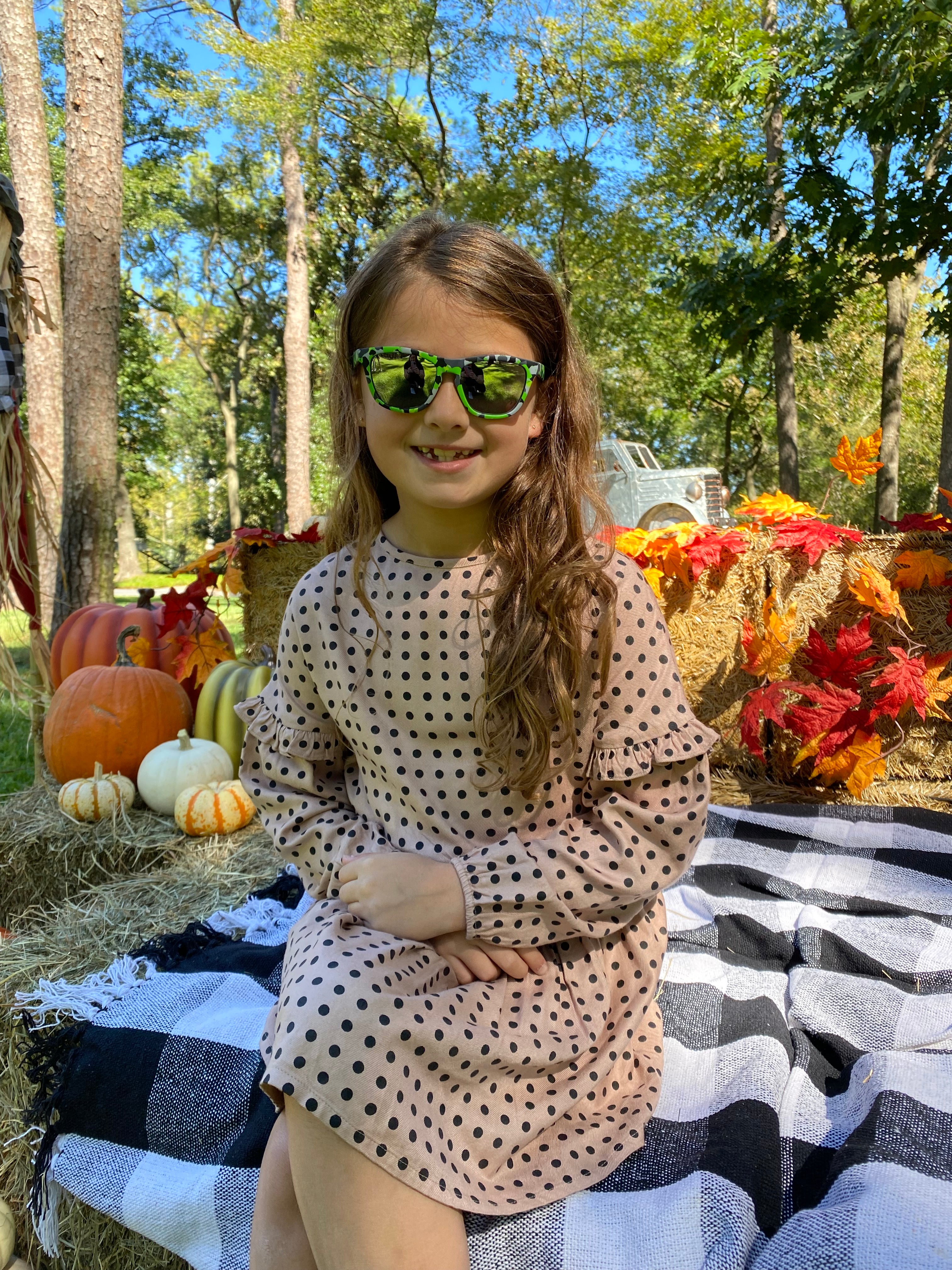 Little girl sitting in a fall dress with pumpkins around wearing polarized kids sunglasses by Sunnies shades in a green camo print with silver reflective lenses.