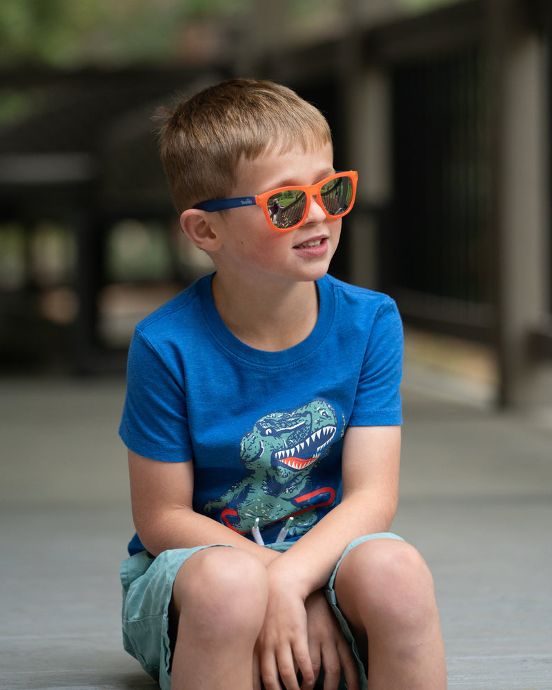 A little boy sitting outside wearing the Milo Man by Sunnies Shades, which are a two tone kids sunglass with a bright orange front, royal blue sides, and silver reflective and polarized lenses.