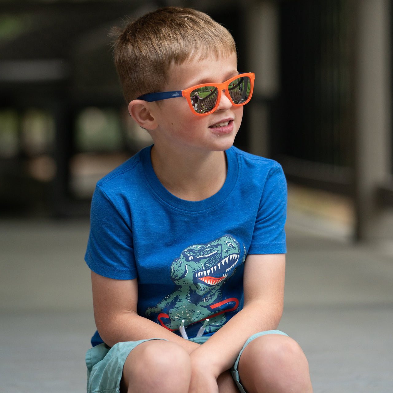 A little boy sitting outside wearing the Milo Man by Sunnies Shades, which are a two tone kids sunglass with a bright orange front, royal blue sides, and silver reflective and polarized lenses.