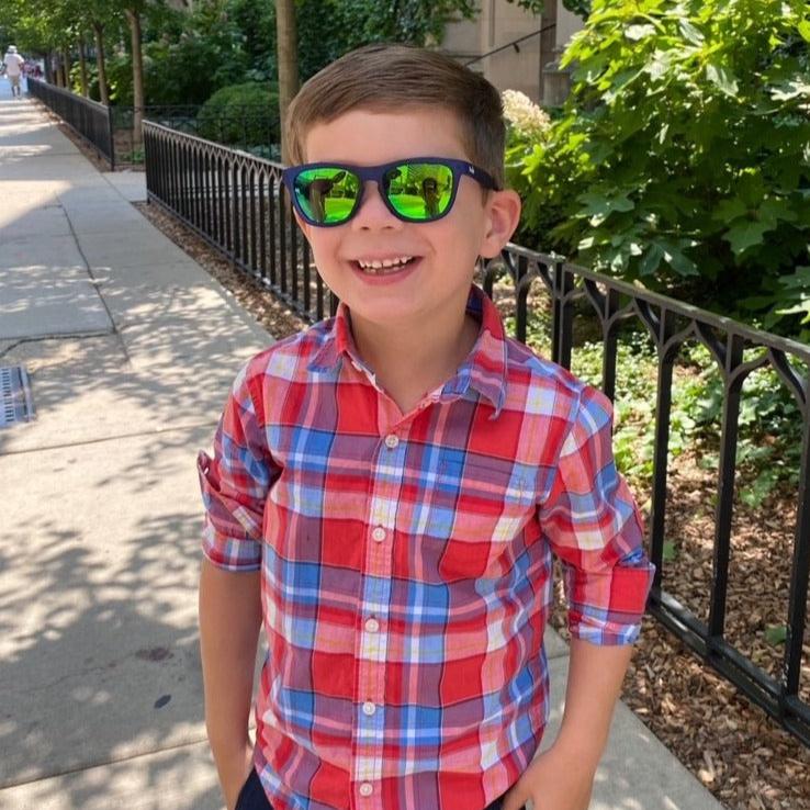 Young boy wearing Thank you Berry Punch polarized kids sunglasses in a transparent blue frame and green reflective lenses.