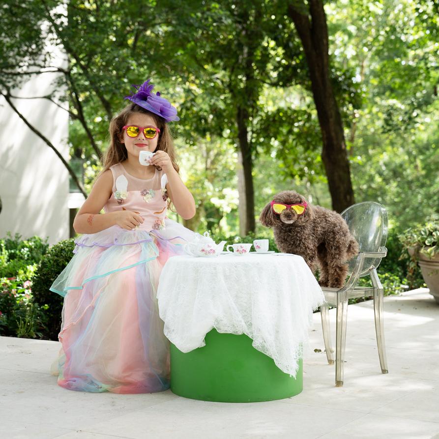 Adorable little girl have tea with a poodle in her polarized kids sunglasses by Sunnies Shades in frame Tea Time with Poodles, which is a transparent pink frame with reflective yellow/green polarized lenses.